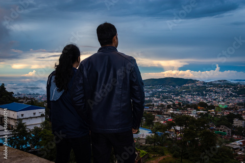 young couple watching downtown city view with dramatic cloudy sky at evening from mountain top