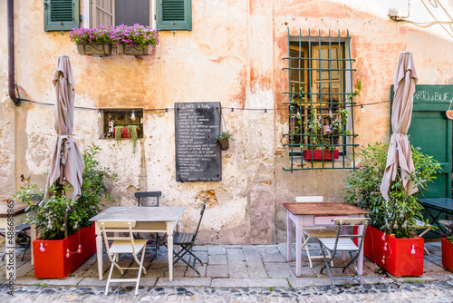 Finalborgo, Finale Ligure, Italy. May 5, 2021. In a small street in the center, two wooden tables outside a restaurant under windows decorated with flowers.