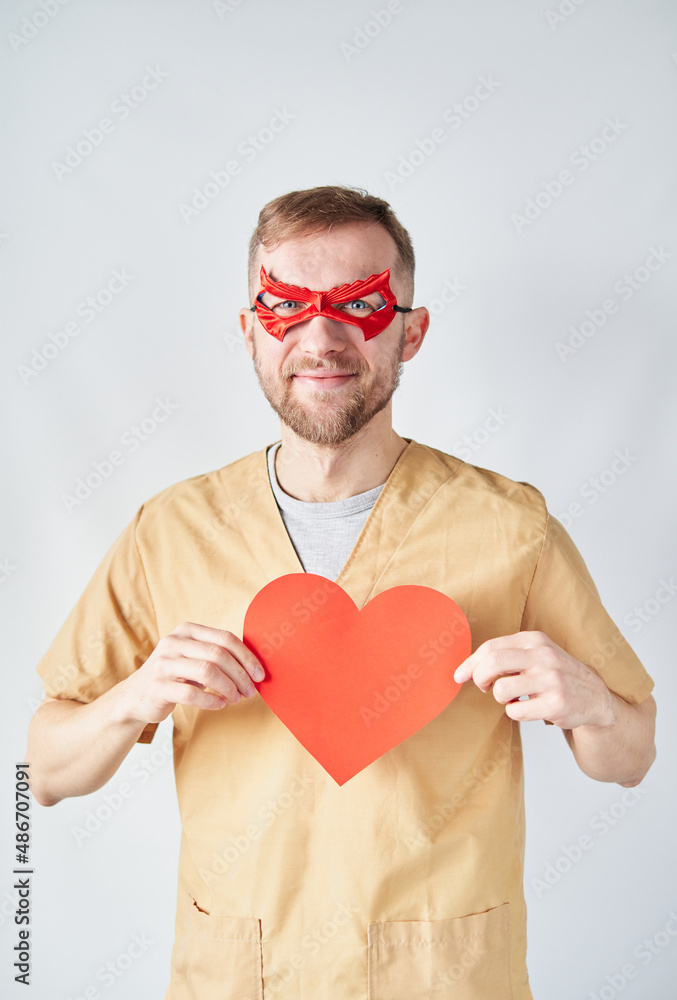 Cheerful caucasian male cardiologist doctor in medical uniform and red superhero mask with red paper heart shape. Adult doctor surgeon in hospital posing over the wall. High quality photo Cheerful caucasian male cardiologist doctor in medical uniform and red superhero mask with red paper heart shape. Adult doctor surgeon in hospital posing over the wall. High quality photo