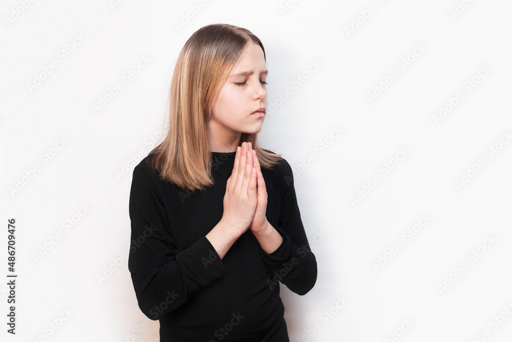 A young girl with a sad, pleading face holds her hands in front of her and prays, talks to God, to herself, her eyes closed. The concept of prayer, peace A young girl with a sad, pleading face holds her hands in front of her and prays, talks to God, to herself, her eyes closed. The concept of prayer, peace
