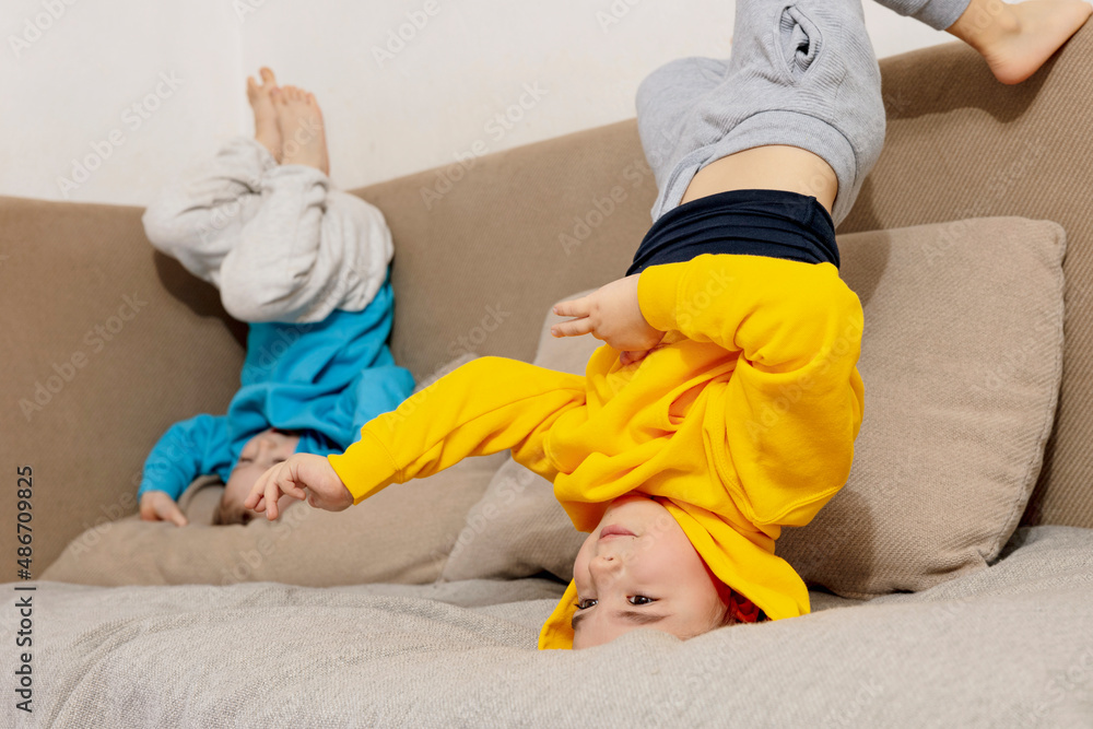 Obraz premium Two little caucasian kids doing gymnastic handstand exercise in living room. Children standing on hands upside down and having fun together. Sport at home. Workout in the apartment. Healthy lifestyle.