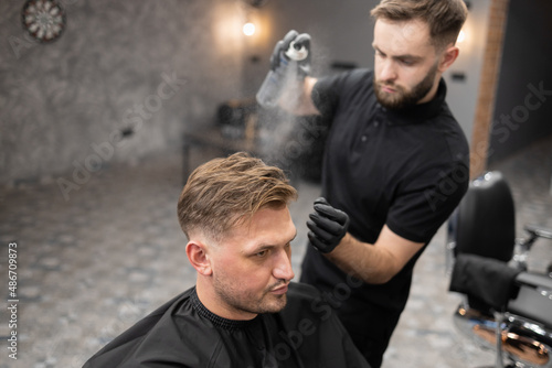 Hairdresser apply hairspray on hair of male client sitting in chair in barbershop.