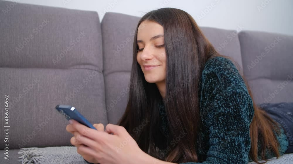 Beautiful young woman dating online and communicating on social media app in modern smartphone. Cheerful brunette female typing message in mobile phone while lying on couch at home with smile on face Beautiful young woman dating online and communicating on social media app in modern smartphone. Cheerful brunette female typing message in mobile phone while lying on couch at home with smile on face