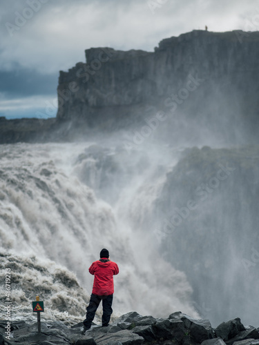 A man in red coat standing at the edge of Dettifoss waterfall, North Iceland. A warning sign to the man's left.