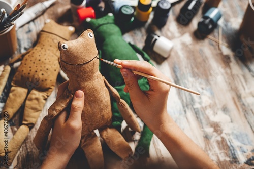 Handmade soft Doll frog. DIY gift making. Human hands hold frog soft toy. Needlework accessories on grey background. Handmade house decor. Process crafting a vintage soft toy. Home workshop. Close-up