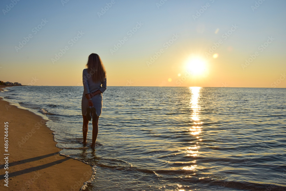 A young woman walks along the sea beach on the shore holding her shoes in her hands
