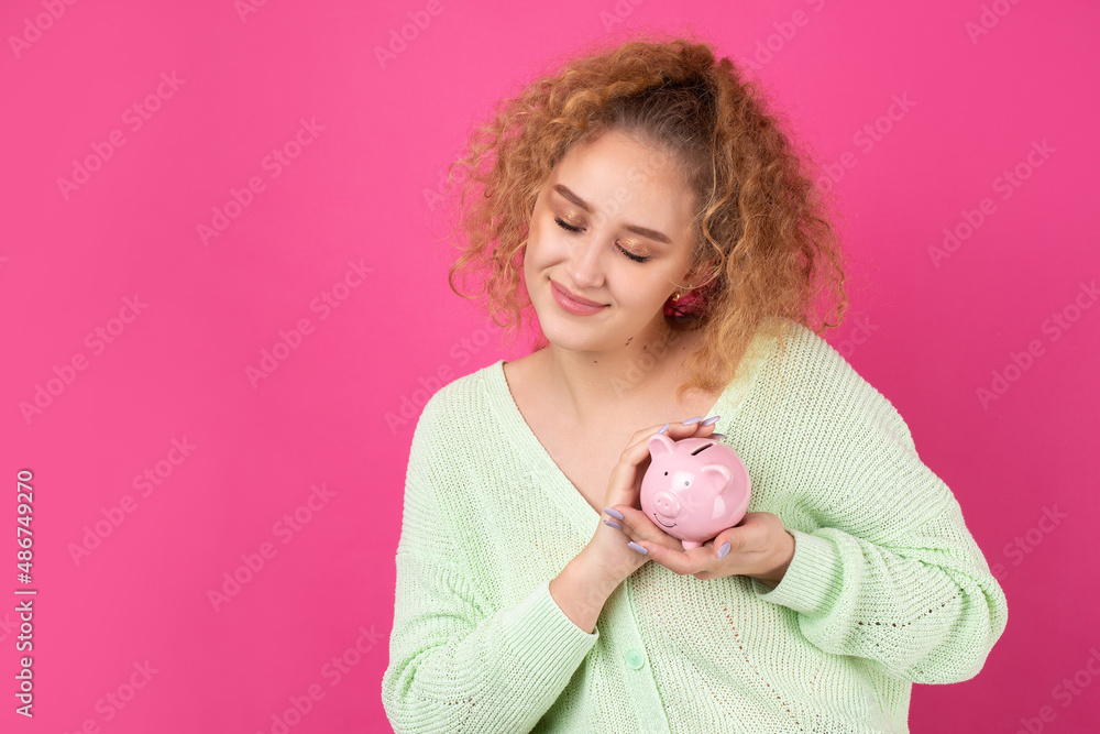 A cute young girl with curly red hair holds a piggy bank, a pink piglet in her hands. The concept of wealth and safety of money. A cute young girl with curly red hair holds a piggy bank, a pink piglet in her hands. The concept of wealth and safety of money.