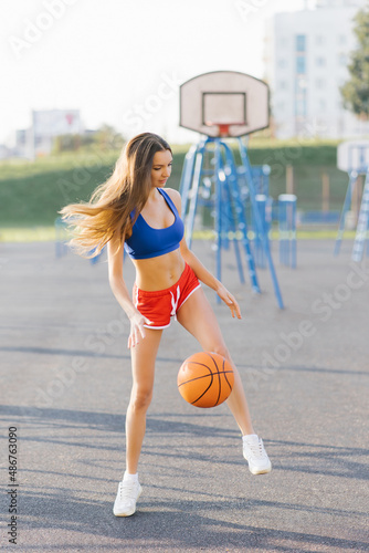 An athletic young woman in sports shorts and T-shirts plays on a basketball court with a ball