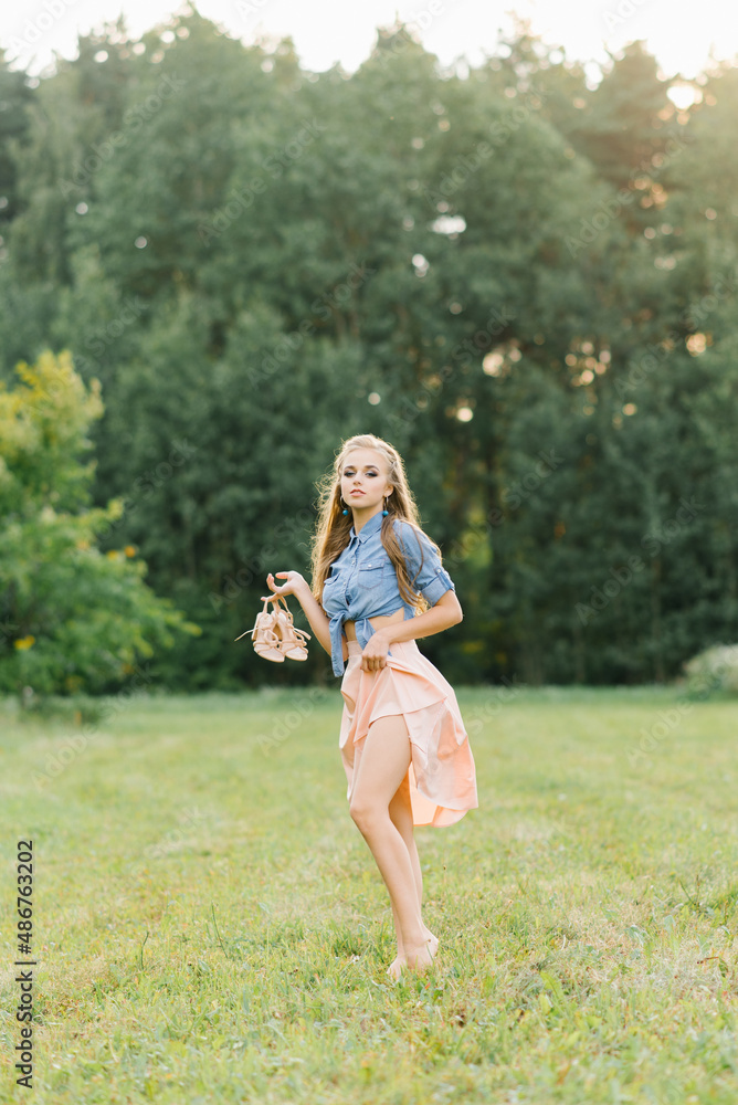 A beautiful young woman holds summer shoes in her hands and walks barefoot on the grass on a summer day