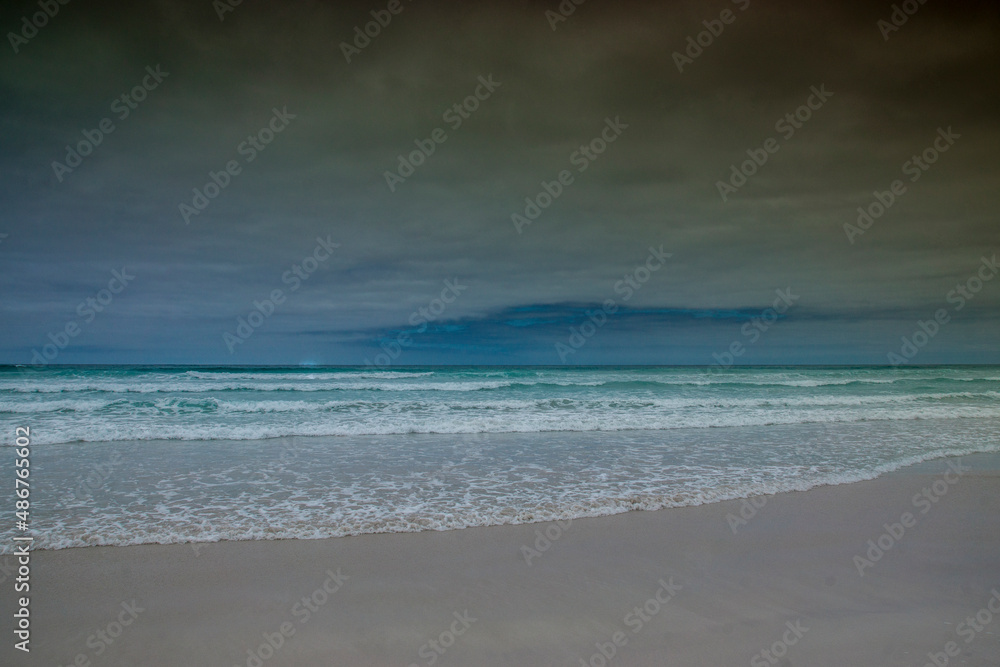 Turquoise, blue and green sea waves on a white sand beach under a grey sky; Tortuga Bay, Galapagos.
