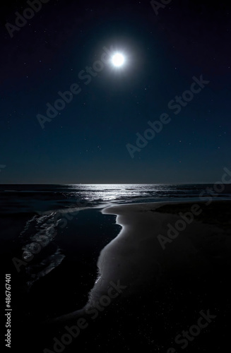 night image of a beach with the moon in the starry sky reflecting on the water of the seashore, painting a cave of light, Isla Canela, Huelva Spain, vertical