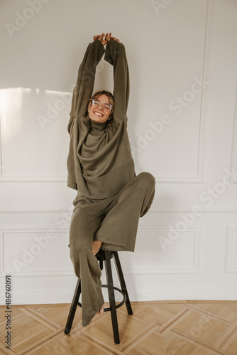 Full length young caucasian woman sits on stool stretching arms up against white background with space for text. Tanned lady wears glasses, grey warm suit with eyes closed. Weekend enjoyment concept