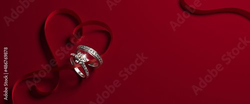 2 white gold engagement rings with diamonds and a heart-shaped ribbon on a red background. Romantic wedding jewelry background.