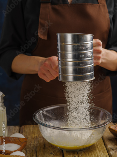 The chef sifts the flour through a sieve into a glass bowl. Flour in frozen flight. The concept is the preparation of dough, recipes for pizza, bread, pasta, confectionery from dough.