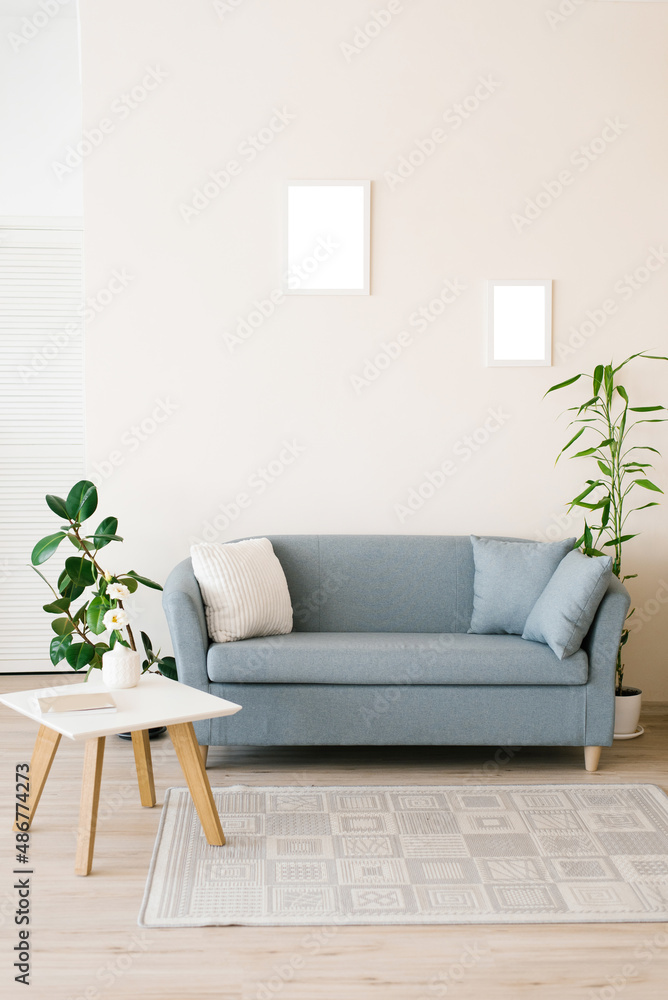 A dusty blue sofa with pillows, a white coffee table, potted plants in a bright cozy living room. Mockup frames for posters on the wall