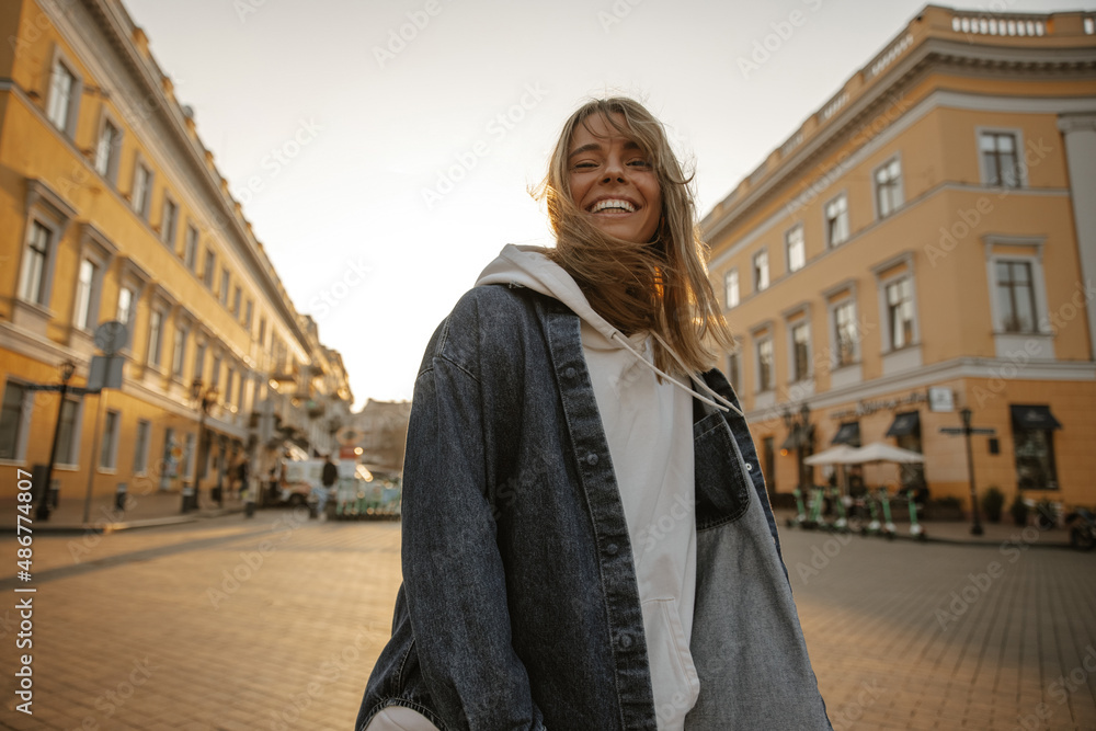 Smiling fashionable young caucasian woman in sporty style posing on street with place for text. Blonde in good mood walks on fresh air. Spring fashion concept