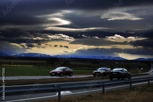 Samochody na drodze szybkiego ruchu, krajobraz wiejski przed zachodem słońca, góry i ciemne chmury. 