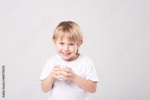Portrait of a blond child with a glass of milk on a white background. Expressive face, smile, eye contact. Vitamins, calcium, milk teeth, dairy products, healthy food, advertising concept. Copy space
