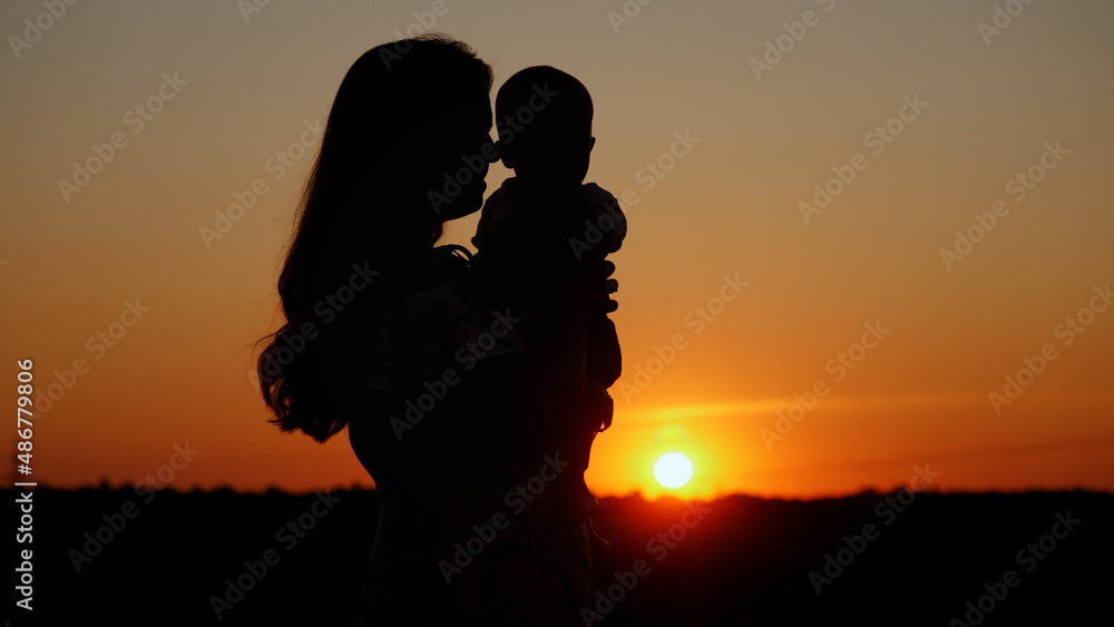 Caring mother holds the baby in her arms and kisses him, unrecognizable silhouette at sunset, motherly love
