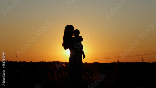 Caring mother holds the baby in her arms and kisses him, unrecognizable silhouette at sunset, motherly love