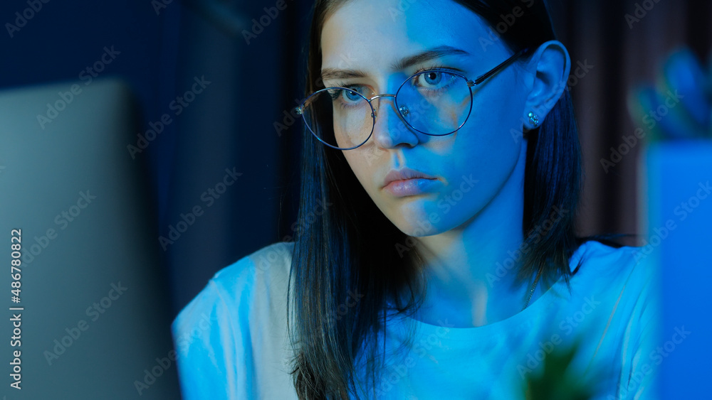 Portrait of teenage girl with glasses looks at the monitor, plays a game late at night or study, room with blue light