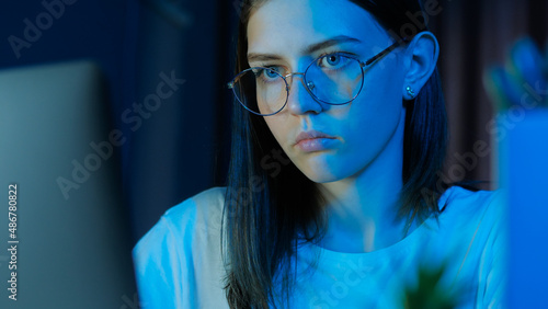 Portrait of teenage girl with glasses looks at the monitor, plays a game late at night or study, room with blue light