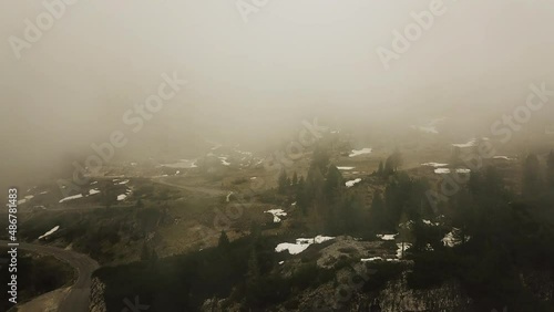 Shooting upward, mysterious mountain landscape disappearing into fog on cold winter day. Aerial view of road among coniferous forest.