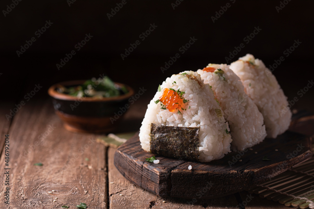 Japanese traditional snack rice balls triangle onigiri with raw salmon and red caviar on a dark wooden board in rustic style Japanese traditional snack rice balls triangle onigiri with raw salmon and red caviar on a dark wooden board in rustic style