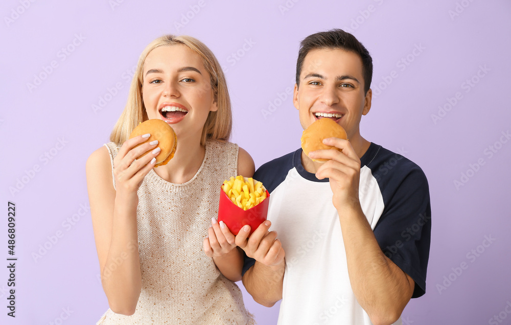 Young couple with french fries eating burgers on purple background Young couple with french fries eating burgers on purple background