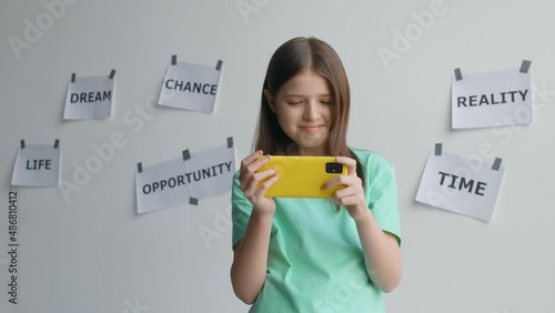 Blonde teenager girl in a T-shirt look into the phone and stands in front of the inscriptions on the wall and smile
