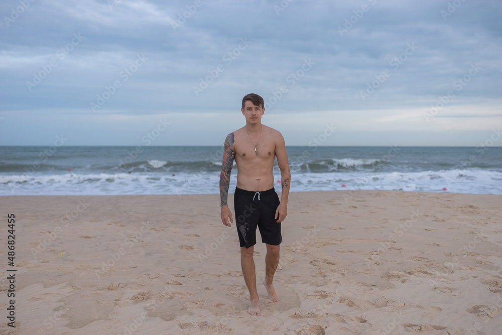 Full height portrait of handsome fit bare-chested young man standing on the beach. guy with tattoo walking by the sea. High quality photo