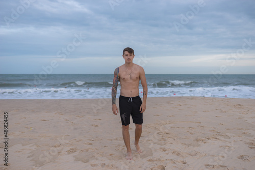 Full height portrait of handsome fit bare-chested young man standing on the beach. guy with tattoo walking by the sea. High quality photo