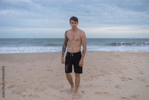 Full height portrait of handsome fit bare-chested young man standing on the beach. guy with tattoo walking by the sea. High quality photo