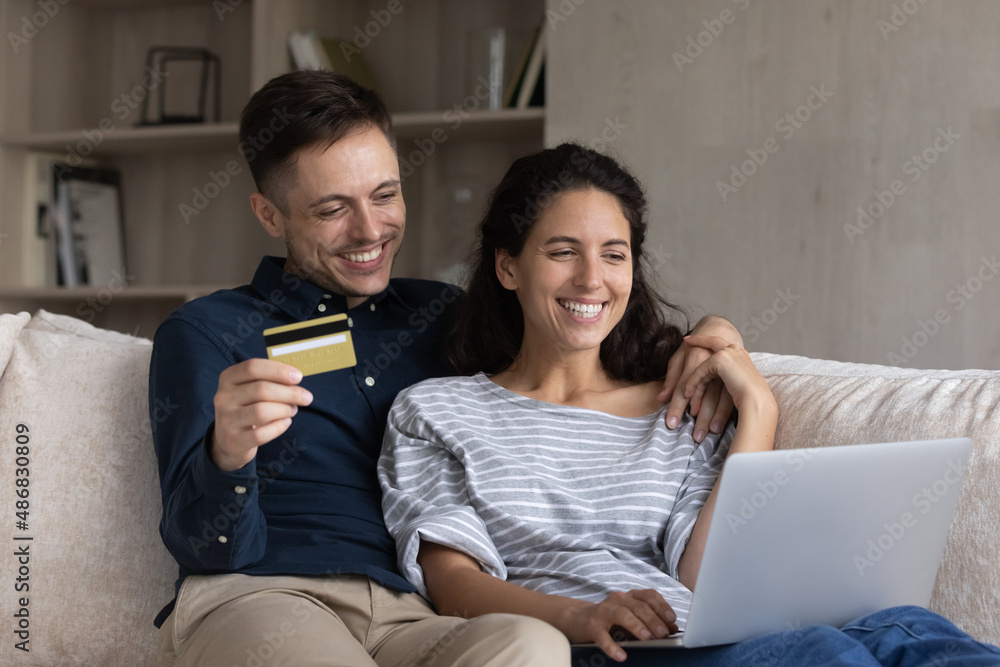 Happy millennial married couple shopping on internet together, using credit card, making payments for purchase from virtual electronic wallet, ordering online, booking hotel on laptop, paying bills