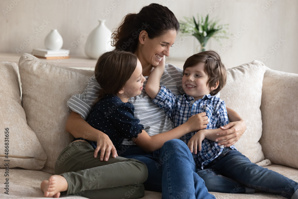 Happy mom hugging little son and daughter with love, care, tenderness, relaxing with kids in couch, talking to children, laughing. Mother and two siblings enjoying leisure at home. Motherhood concept Happy mom hugging little son and daughter with love, care, tenderness, relaxing with kids in couch, talking to children, laughing. Mother and two siblings enjoying leisure at home. Motherhood concept