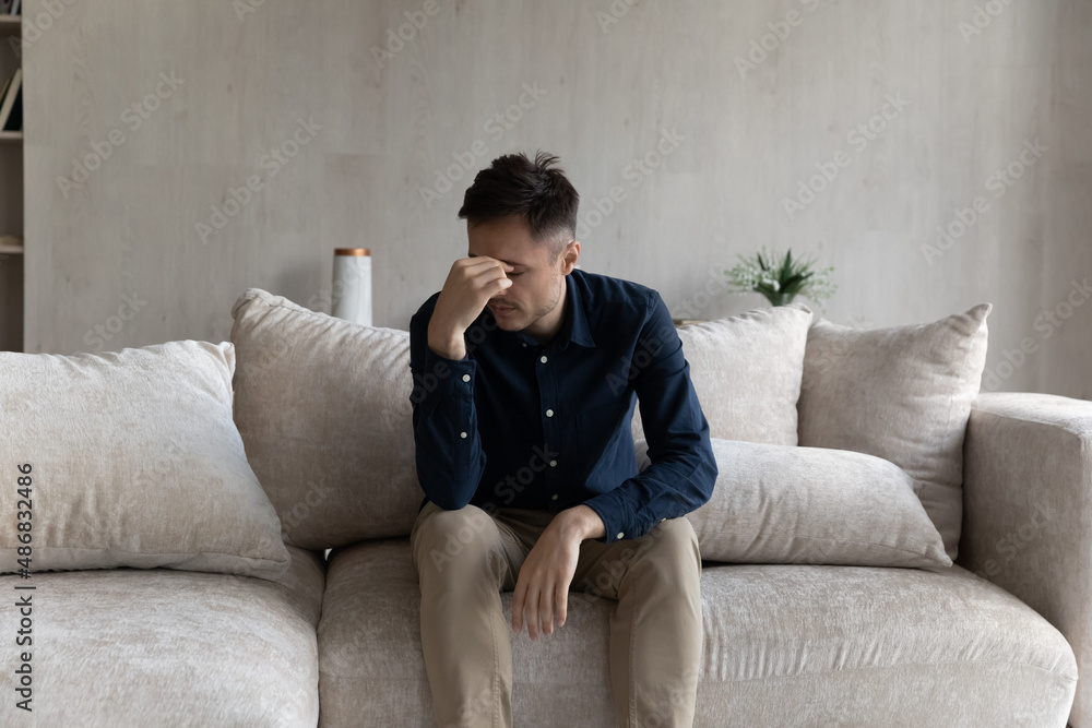 Tired frustrated young man feeling exhausted, suffering from migraine, sitting on cough at home, touching head, eyes, going through stress, apathy, grief, loss. Depression, fatigue concept Tired frustrated young man feeling exhausted, suffering from migraine, sitting on cough at home, touching head, eyes, going through stress, apathy, grief, loss. Depression, fatigue concept