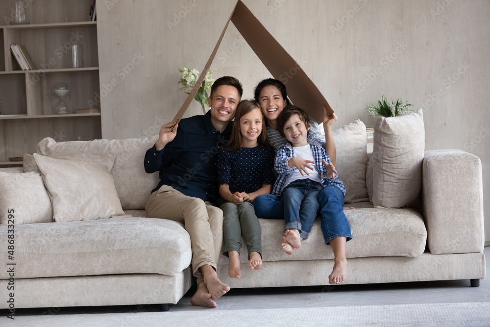 Happy young parents and two cute kids holding cardboard paper roof of house overhead, showing symbol of safe home, protection, insurance, relocation, looking at camera, smiling. Wide shot portrait Happy young parents and two cute kids holding cardboard paper roof of house overhead, showing symbol of safe home, protection, insurance, relocation, looking at camera, smiling. Wide shot portrait