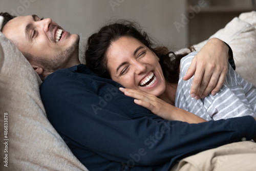 Joyful millennial couple having fun and enjoying closeness at home, hugging on couch, relaxing, talking, laughing, showing perfect teeth. Happy husband and wife resting on soft comfortable couch