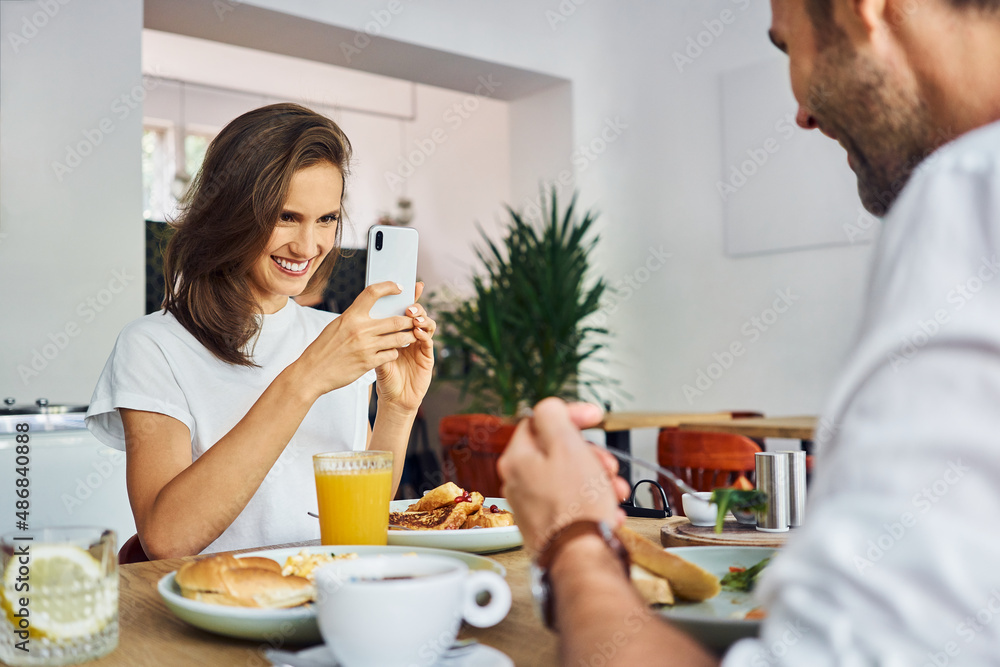 Woman photographing her friend at cafe using smartphone while eating breakfast together