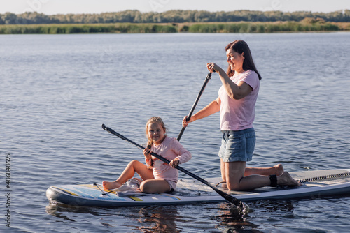 Middle aged mother teaching little female kid to do puddle boarding on lake with green trees and reeds in background. Active lifestyle. Teaching children to love sports from early age. 