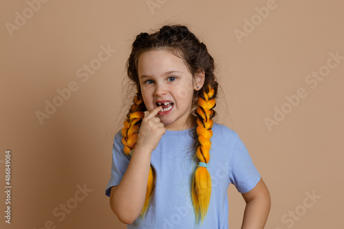 Little, Caucasian girl putting fingers in mouth showing missing tooth looking at camera having kanekalon braids on head on beige background in light blue t-shirt.