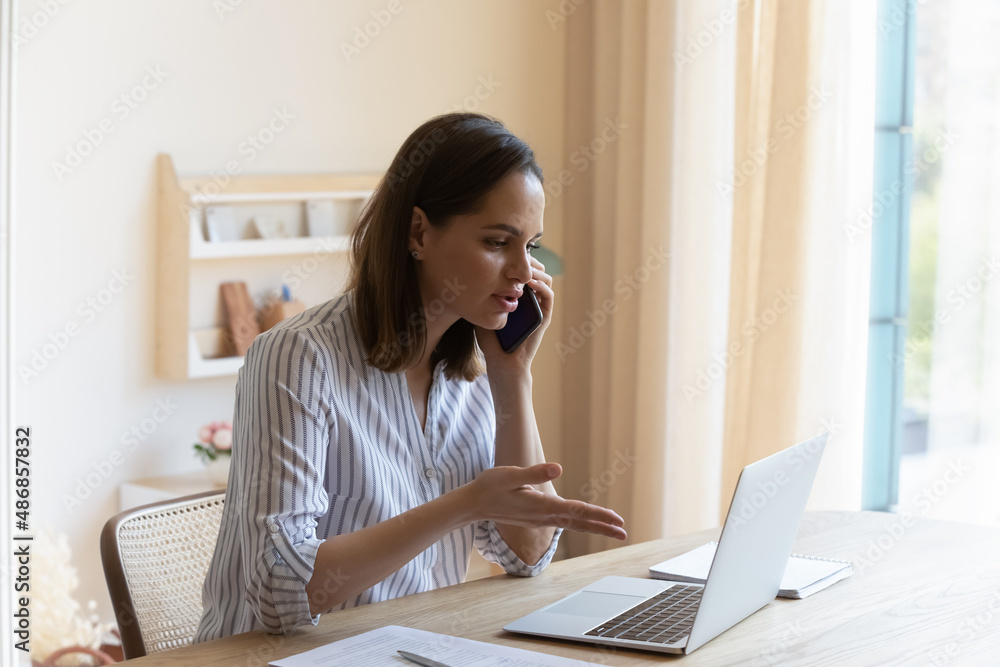 Focused serious young business woman manager entrepreneur looking at laptop screen, communicating by cellphone call with client, discussing problems solution or arguing, multitasking in office. Focused serious young business woman manager entrepreneur looking at laptop screen, communicating by cellphone call with client, discussing problems solution or arguing, multitasking in office.