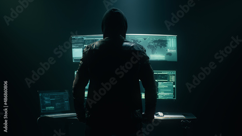 Back view of unrecognizable man in hoodie standing near desk and reading stolen data from computer, monitors in dark room before massive cyber attack on servers. photo