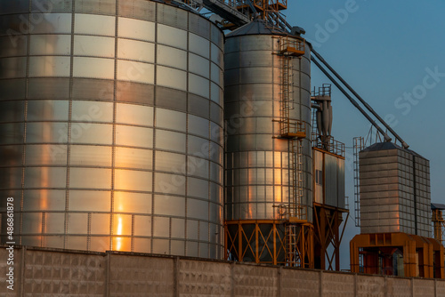 A large modern plant located near a wheat field for the storage and processing of grain crops. view of the granary illuminated by the light of the setting sun against the blue sky. harvest season.