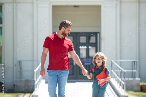 Parent and pupil of primary school go hand in hand. Teacher in t-shirt and cute schoolboy with backpack near school park. photo