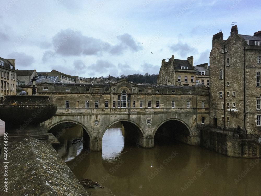 Pulteney bridge over Avon river in Bath. Somerset, England. Old town, stone walls, picturesque spot, blue sky, flying bird, dark water. Chimney. Rapids on a river. Stream. Moss