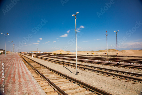 Abstract view of railroad platform and station in desert. Paving platform left, electric lighting masts, loudspeakers on poles. Rails with wooden sleepers.