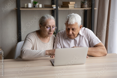 Focused mature wife teaching older husband to use online banking app on laptop. Elder couple using computer at table, browsing internet, shopping, making payment, virtual order, video call