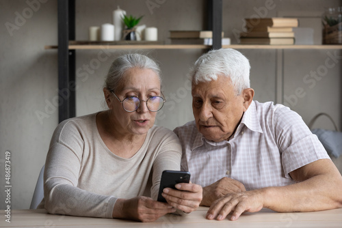 Focused old grandparents couple sharing mobile phone, using virtual ecommerce banking service for shopping, payment, online order. Mature wife explaining elder husband internet app work