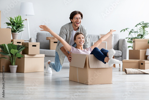 Satisfied caucasian millennial guy pushes wife in box in room interior, have fun together enjoy moving to new home